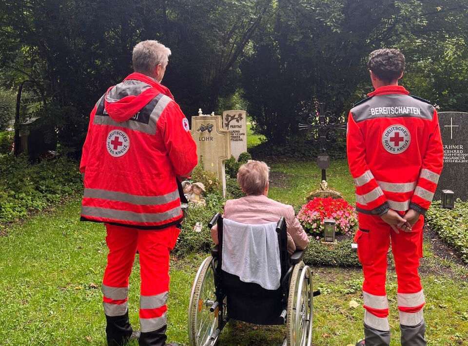 Zwei Rotkreuzler stehen neben einer Seniorin im Rollstuhl vor einem Grab auf einem Friedhof