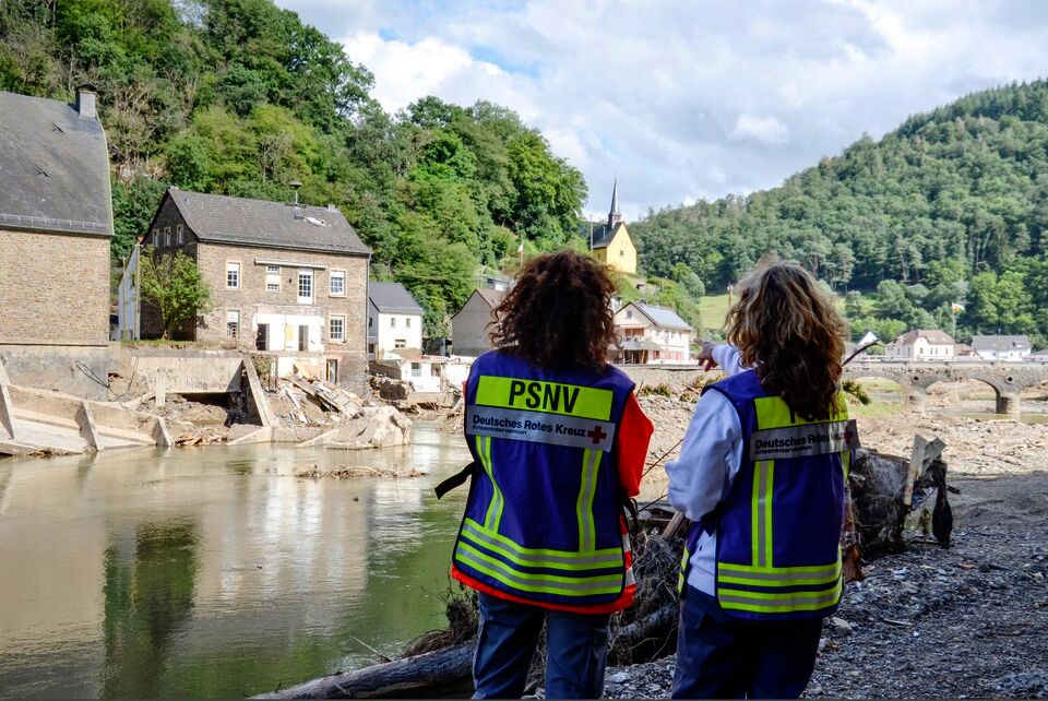 Zwei Helferinnen mit Kennzeichnungswesten mit der Aufschrift PSNV stehen 2021 am Ufer der Ahr und blicken auf die Zerstörung durch das Hochwasser